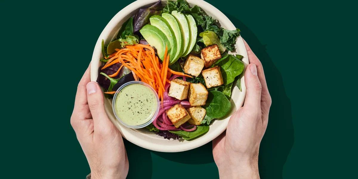 Two hands holding a bowl of salad with avocado, tofu and carrot