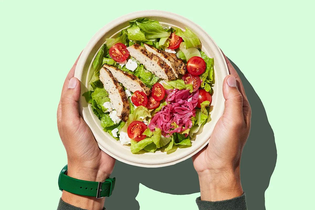 Two hands holding a bowl of chicken salad with tomato and pickled onion