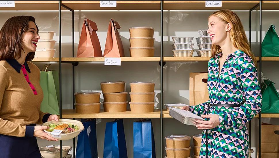 Two women laughing together beside a shelf of packed lunches in paper bags and bowls
