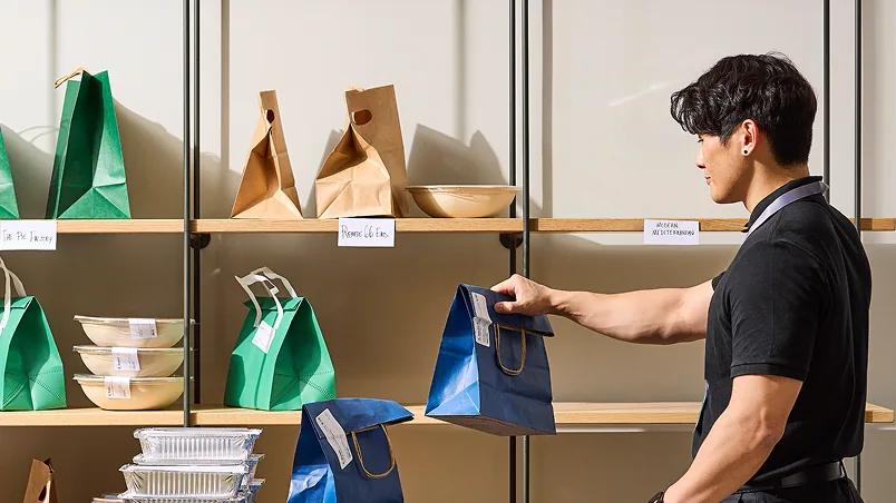 A man in a black shirt placing a bagged lunch on a shelf beside a cart of packed orders