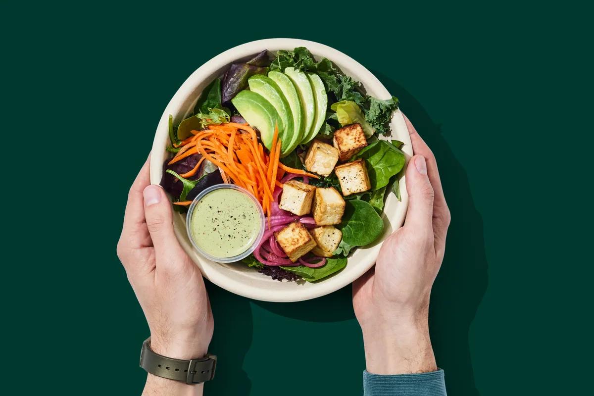 Two hands holding a bowl of salad with avocado, tofu and carrot