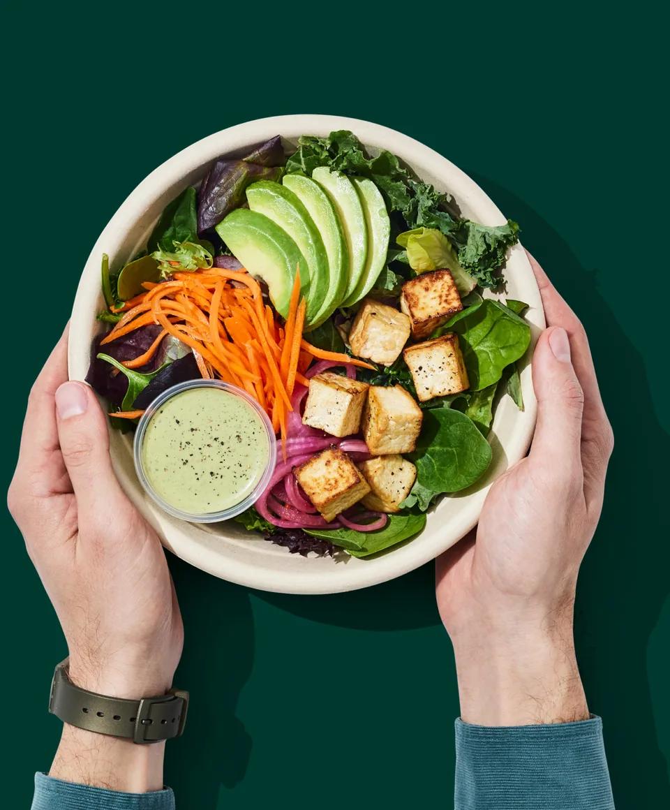Two hands holding a bowl of salad with avocado, tofu and carrot