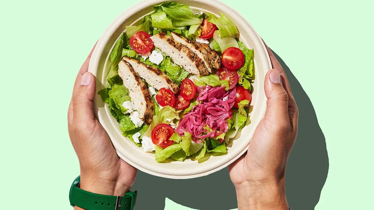 Two hands holding a bowl of chicken salad with tomato and pickled onion