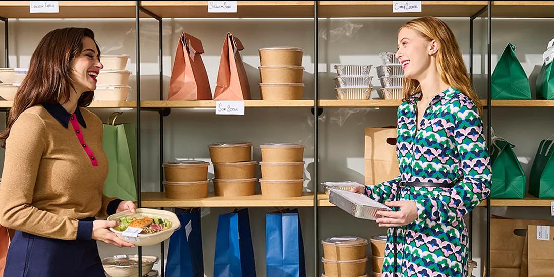 Two women laughing together beside a shelf of packed lunches in paper bags and bowls