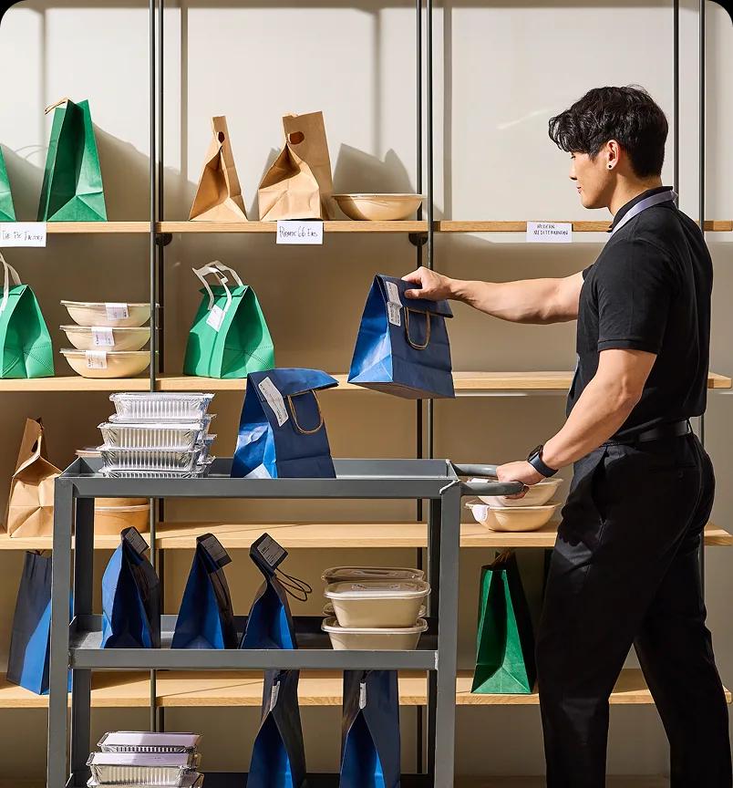 A man in a black shirt placing a bagged lunch on a shelf beside a cart of packed orders