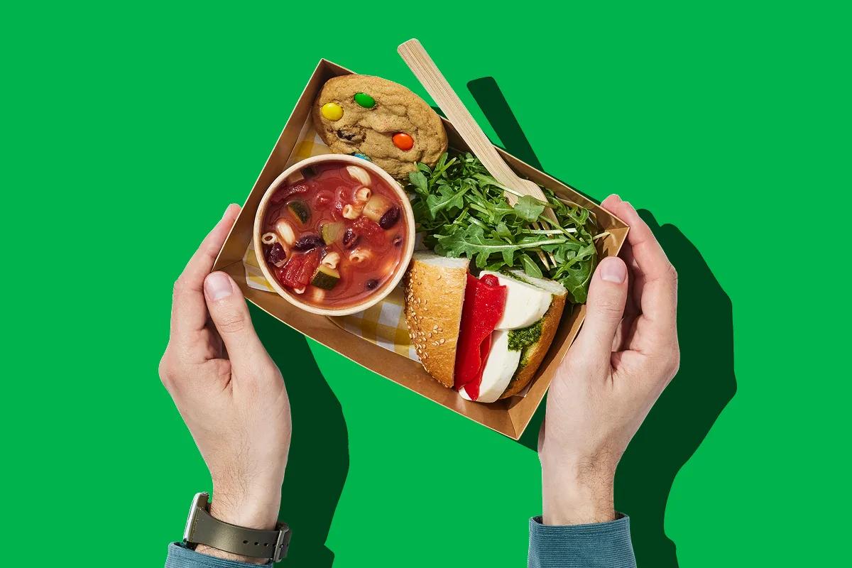 Two hands holding a tray with soup, a sandwich and a cookie