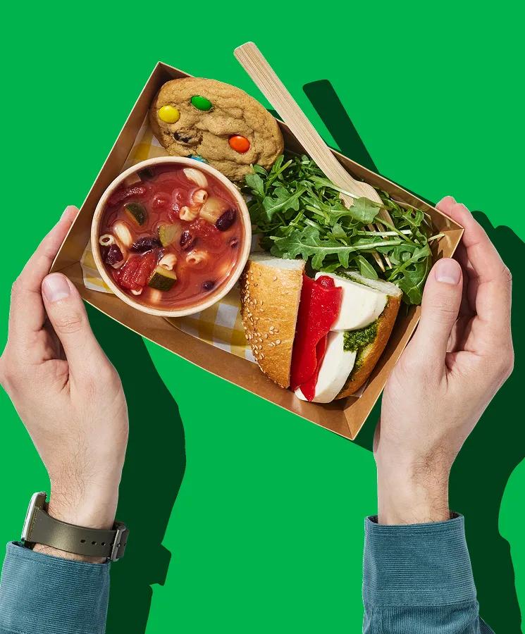 Two hands holding a tray with soup, a sandwich and a cookie